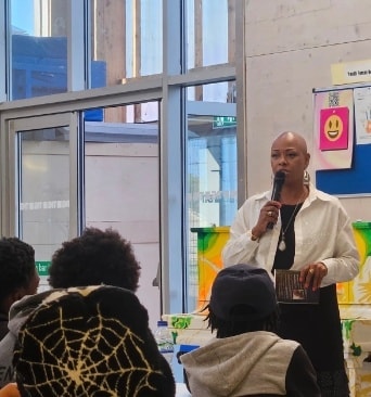 Sharon-Ann Phillips speaking to a group of young people in a community space, holding a microphone and her book